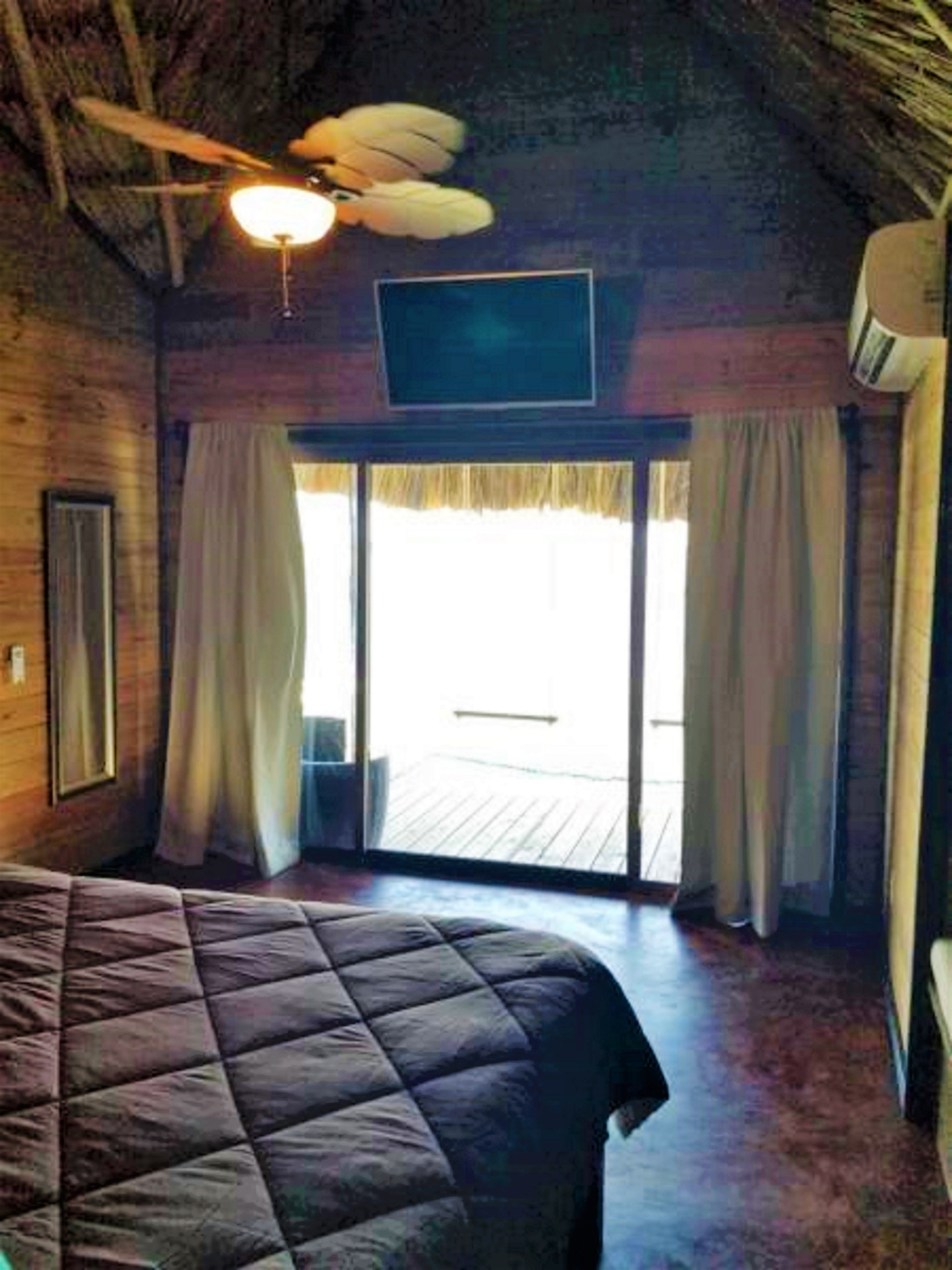 Habitación con cama, ventilador de techo, televisor y puerta corrediza de vidrio que da a un espacio exterior.