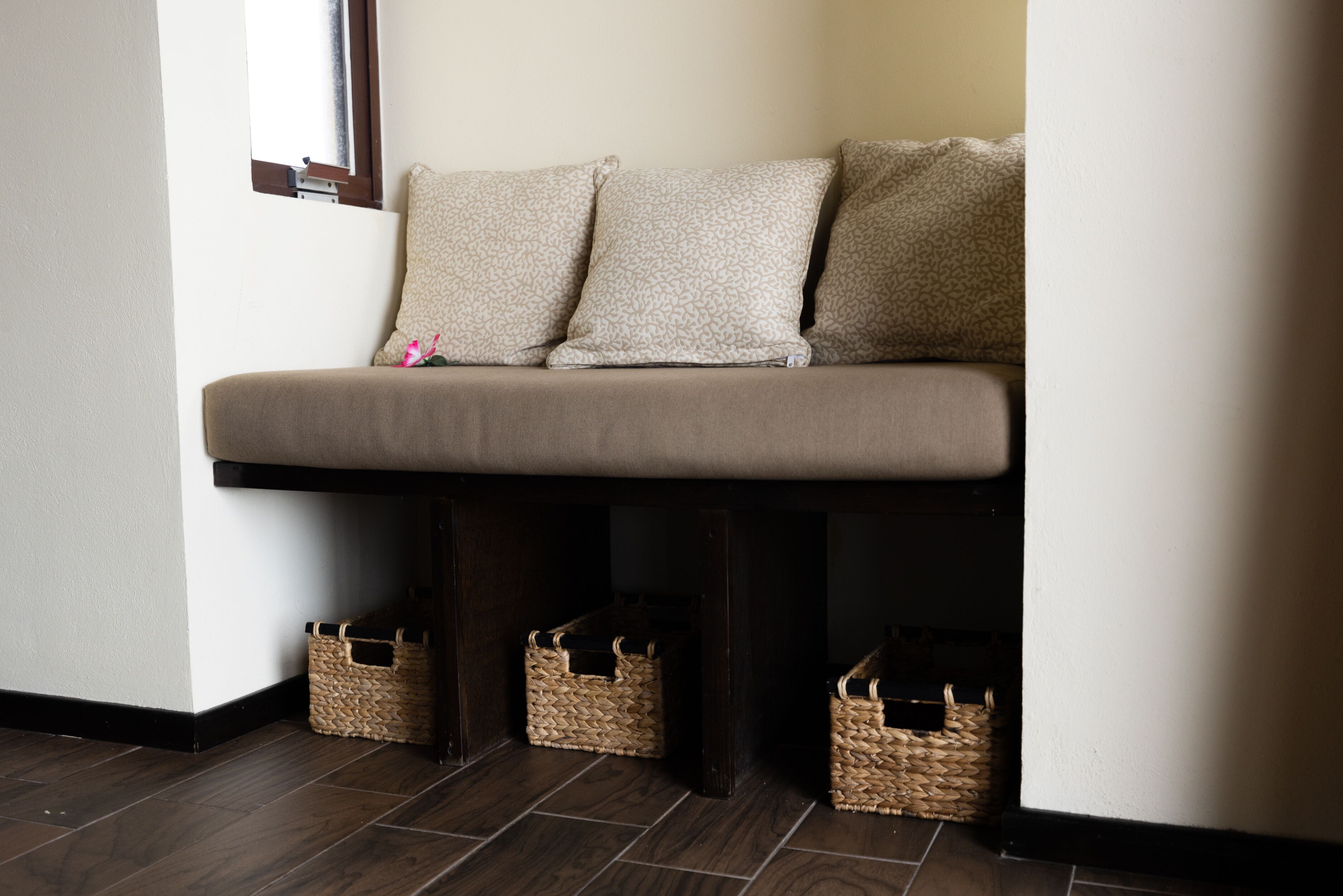 A bench with three pillows, two baskets under it, and a window to the left.