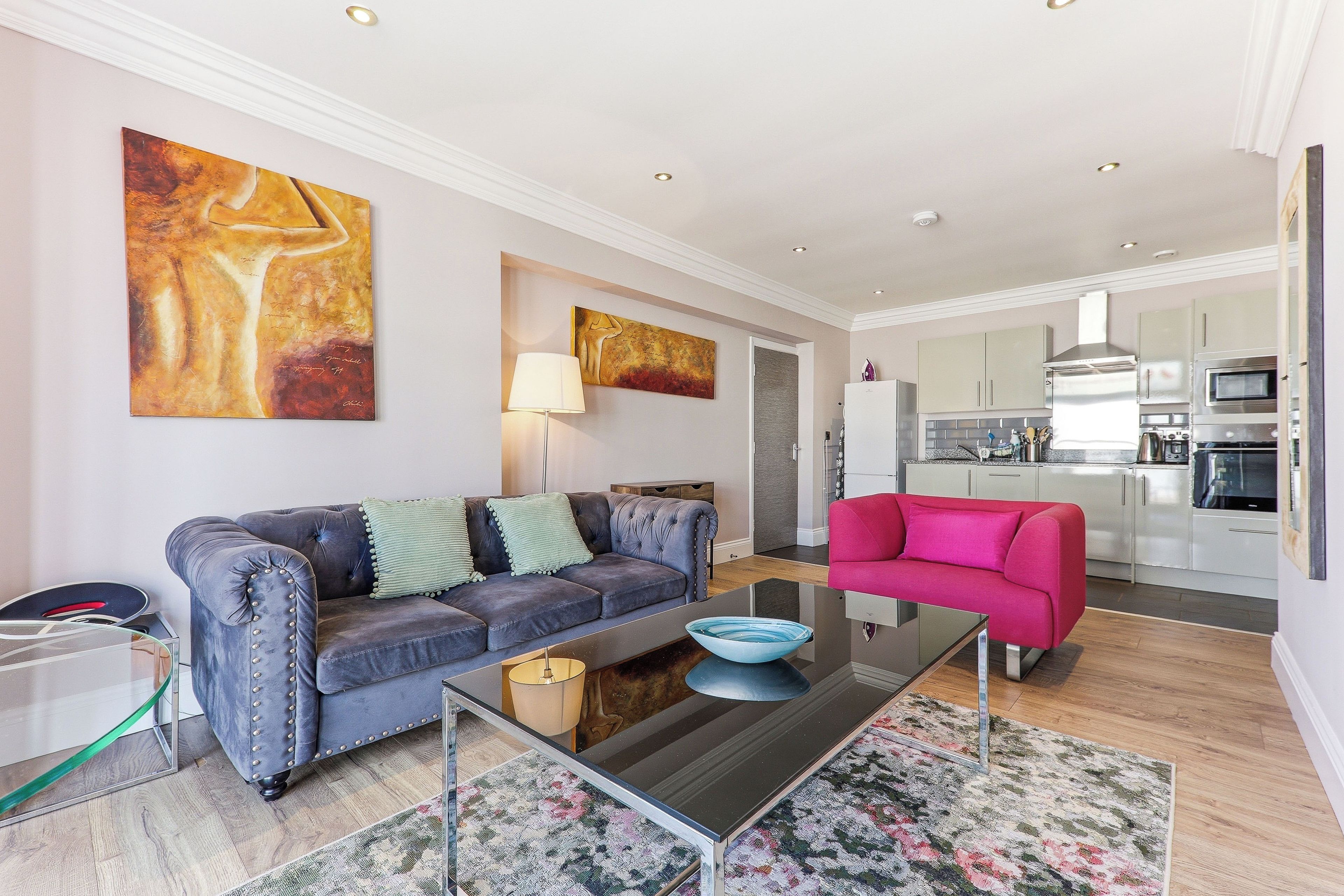 A modern living room with a sofa, a coffee table, and a pink chair. There is a kitchen visible in the background with modern appliances.