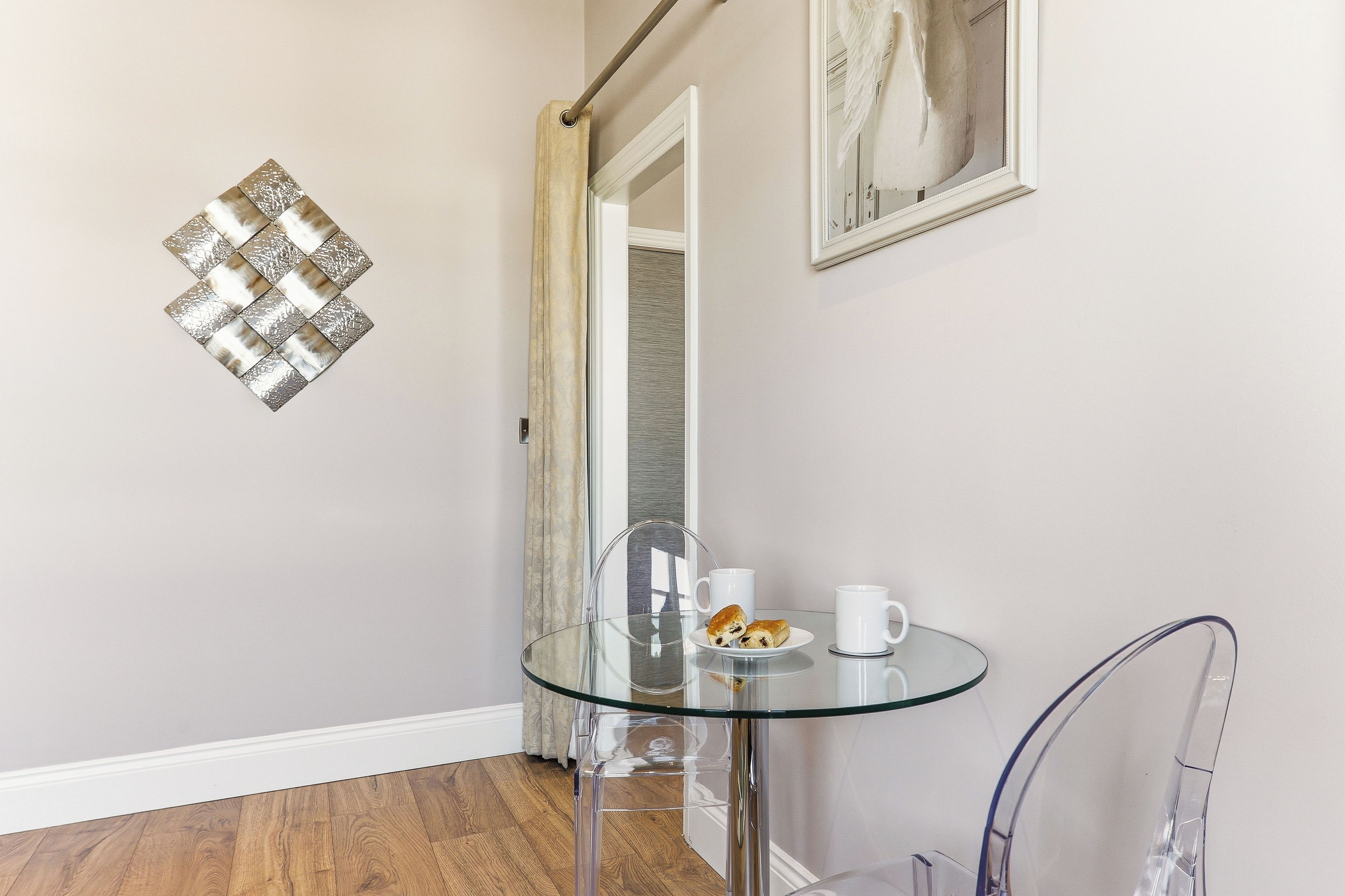 A glass-top table with a plate of pastries and two white cups, a clear chair, a decorative wall piece, and a mirror reflecting a door.