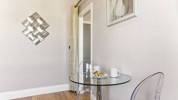 A glass-top table with a plate of pastries and two white cups, a clear chair, a decorative wall piece, and a mirror reflecting a door.
