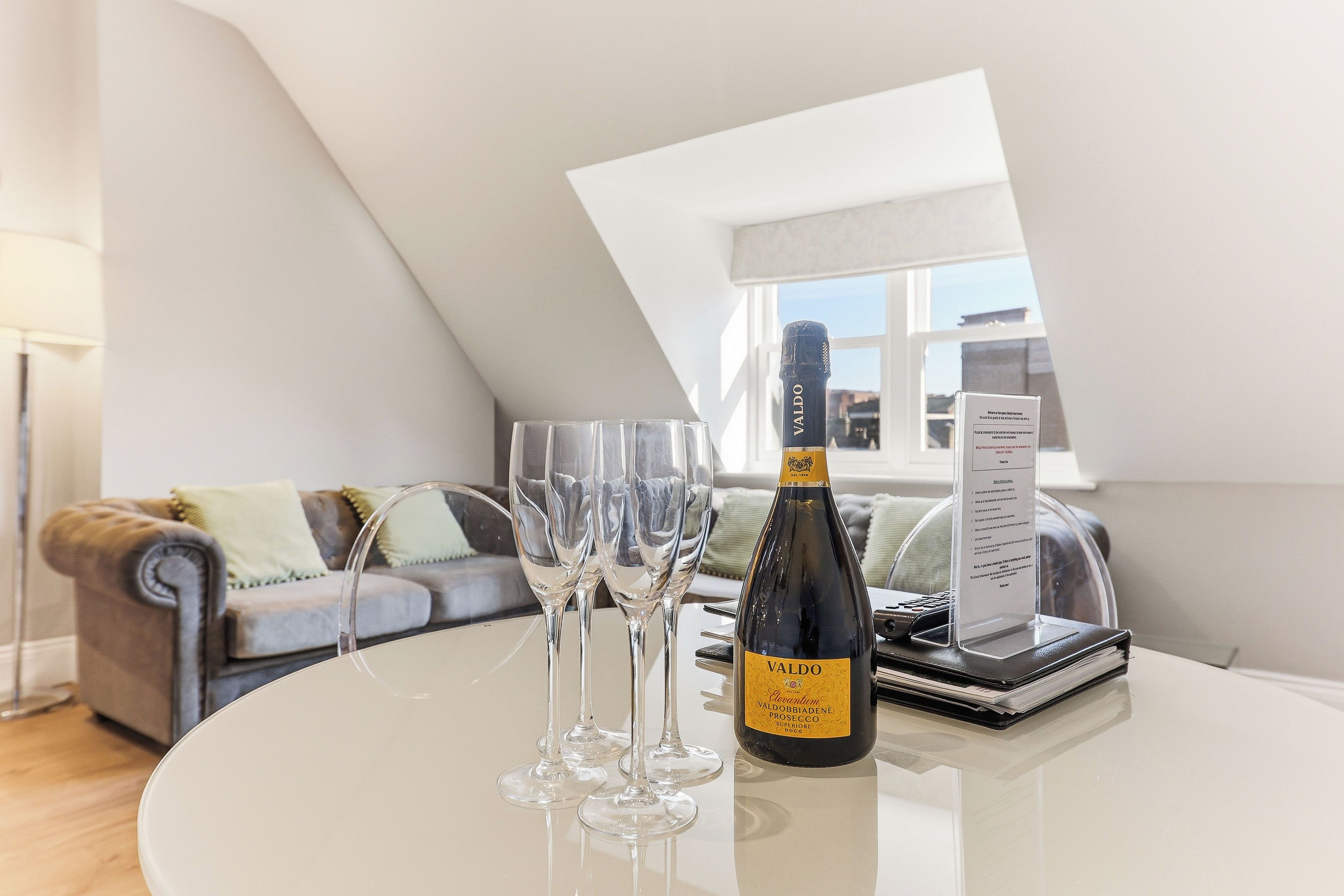 A bottle of Valdo wine on a table with three glasses, a menu, and a laptop in a modern living room with a sloped ceiling and a view through a window.