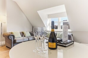 Una botella de vino Valdo sobre una mesa con tres copas, un menú y una computadora portátil en un salón moderno con techo inclinado y vista por una ventana.