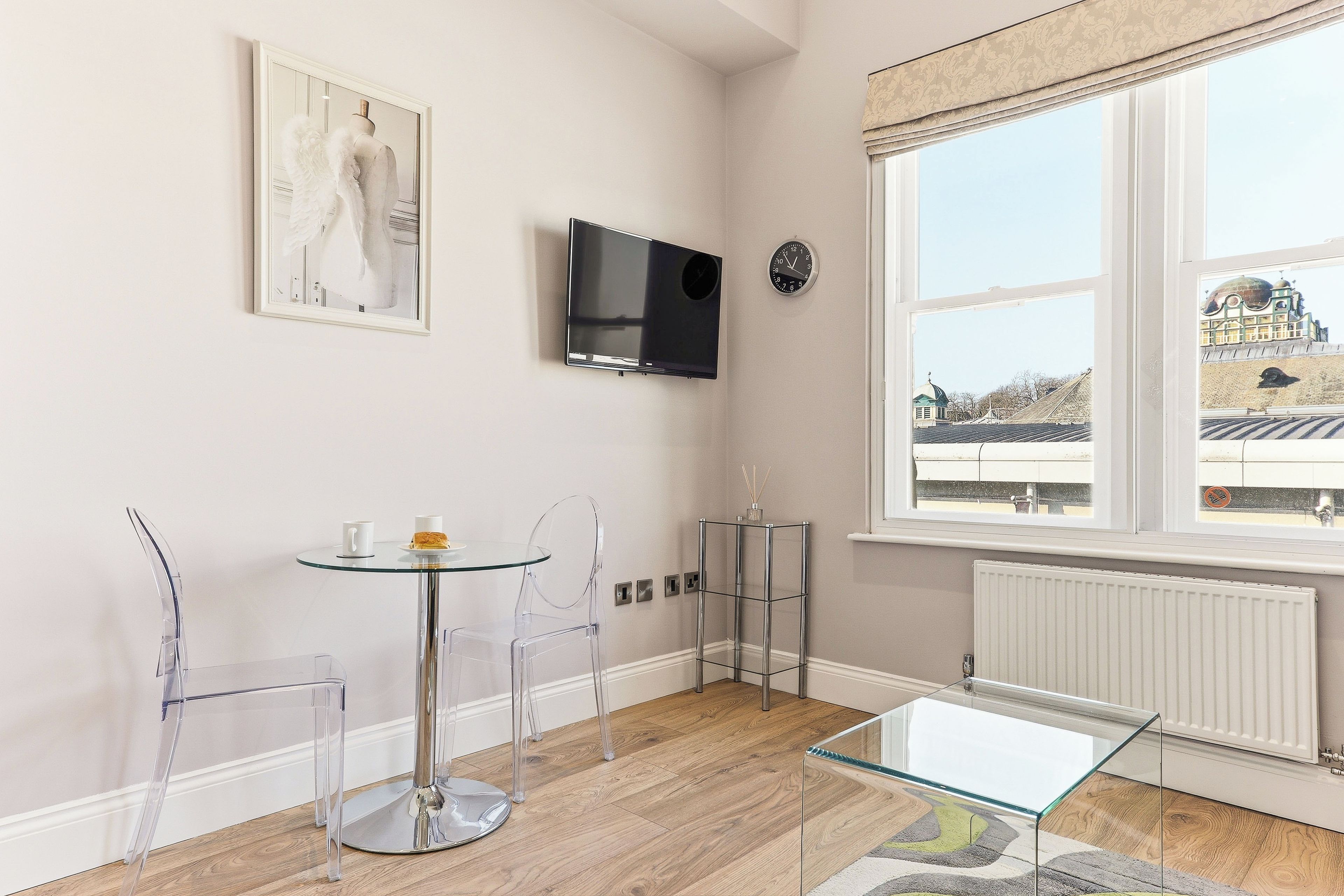 A modern room with a glass table, clear chairs, a mounted TV, a clock, and a window with a view of a building.