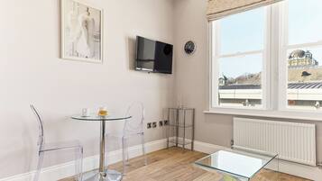 A modern room with a glass table, clear chairs, a mounted TV, a clock, and a window with a view of a building.