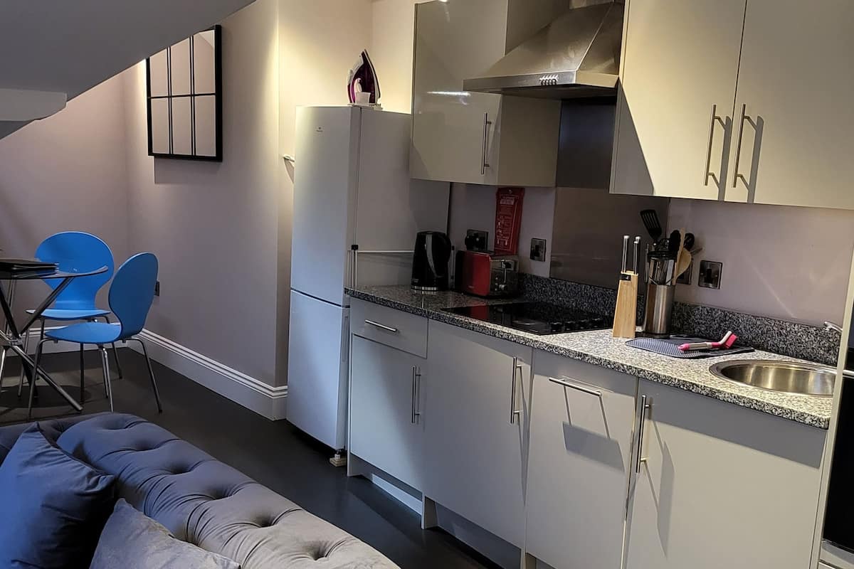 A modern kitchen with grey cabinets, a stainless steel sink, and a built-in oven. There is a dining area with blue chairs and a grey sofa.