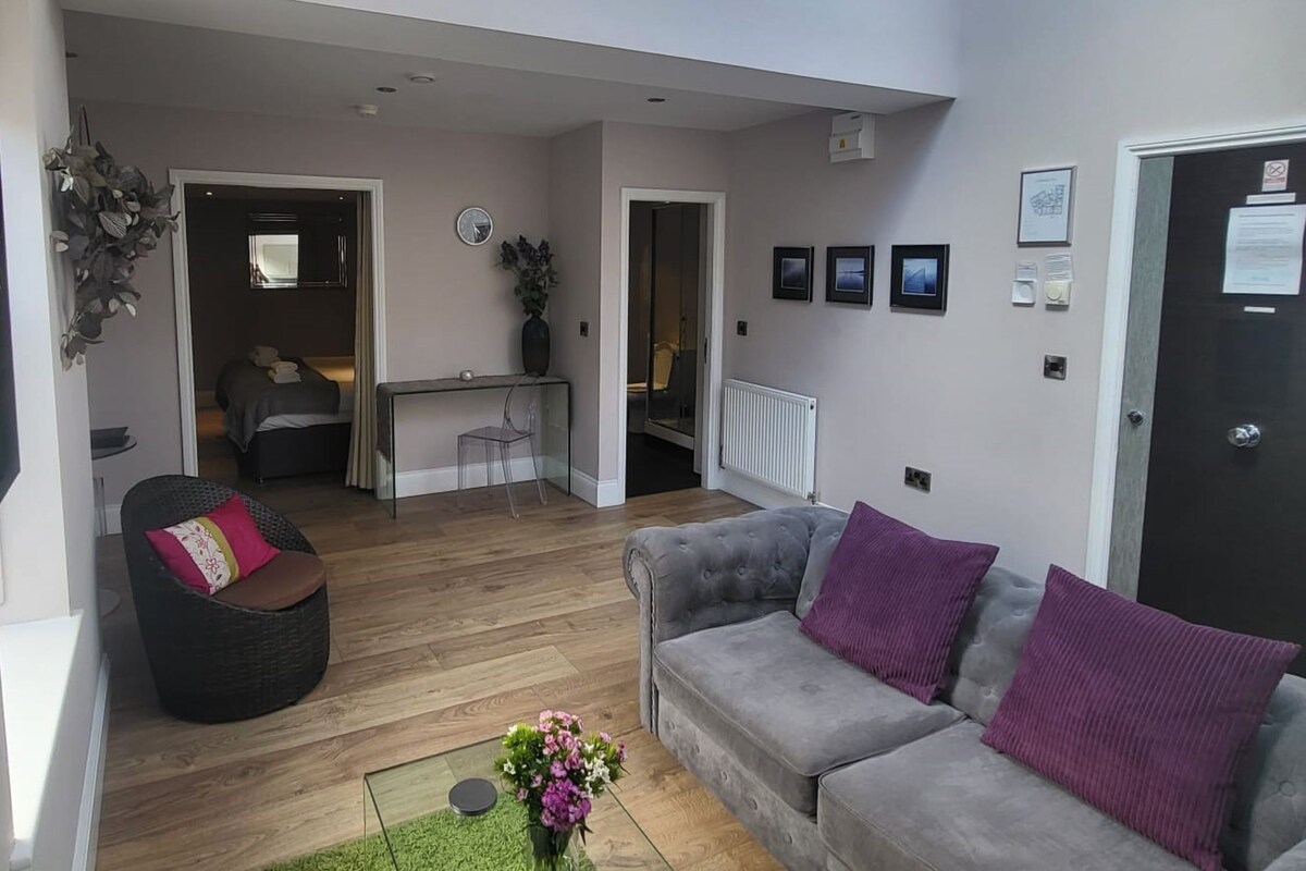 A modern living room with a grey sofa, a black chair, and a small table. There is a wooden floor and a green rug.