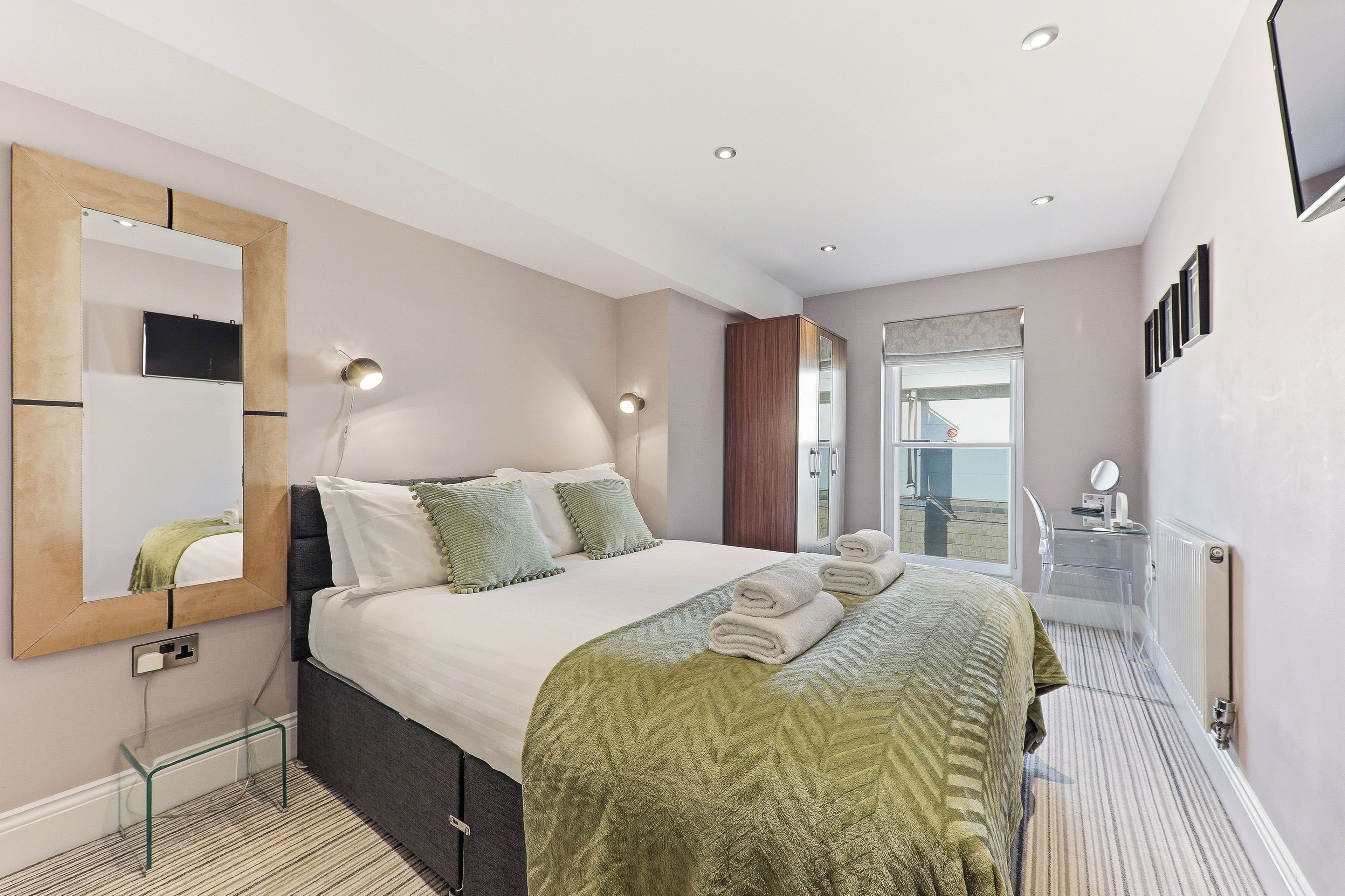 A hotel room with a bed, a mirror, a bedside table, a radiator, and a window.