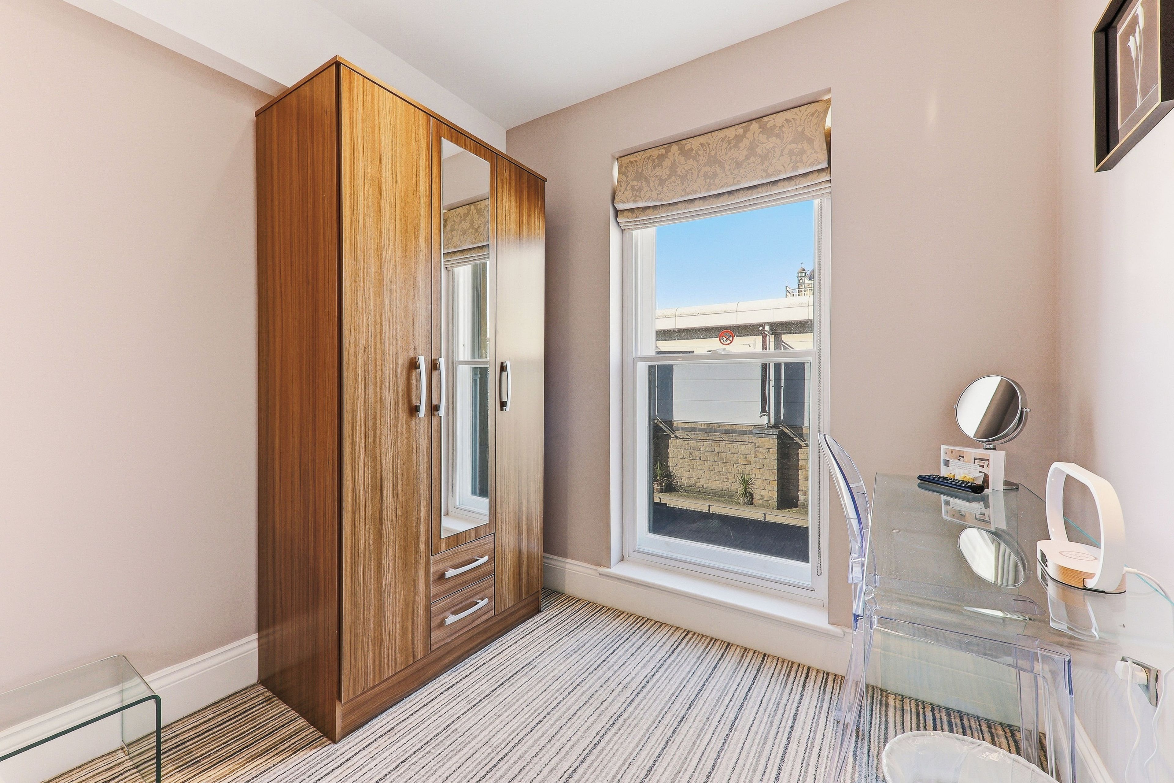 A room with a wooden wardrobe, a window with a view, a glass table with a mirror, and a striped rug.