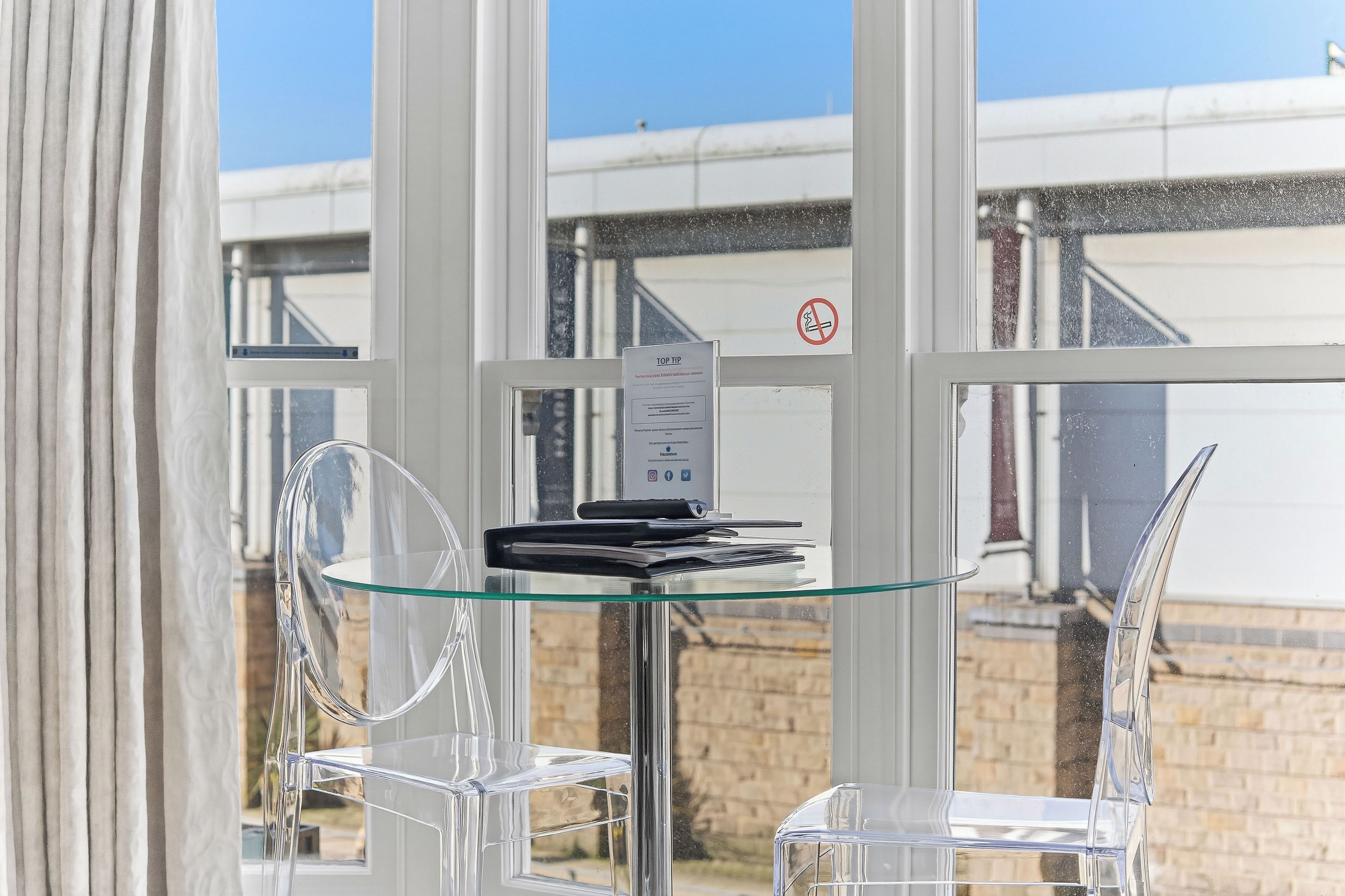 Een transparante glazen tafel met een zwart apparaat, mogelijk een tablet of een klein beeldscherm, voor een raam met uitzicht op een gebouw.