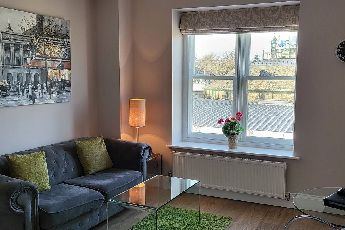A modern living room with a sofa, a glass coffee table, a lamp, a window with a view of buildings, and a clock on the wall.