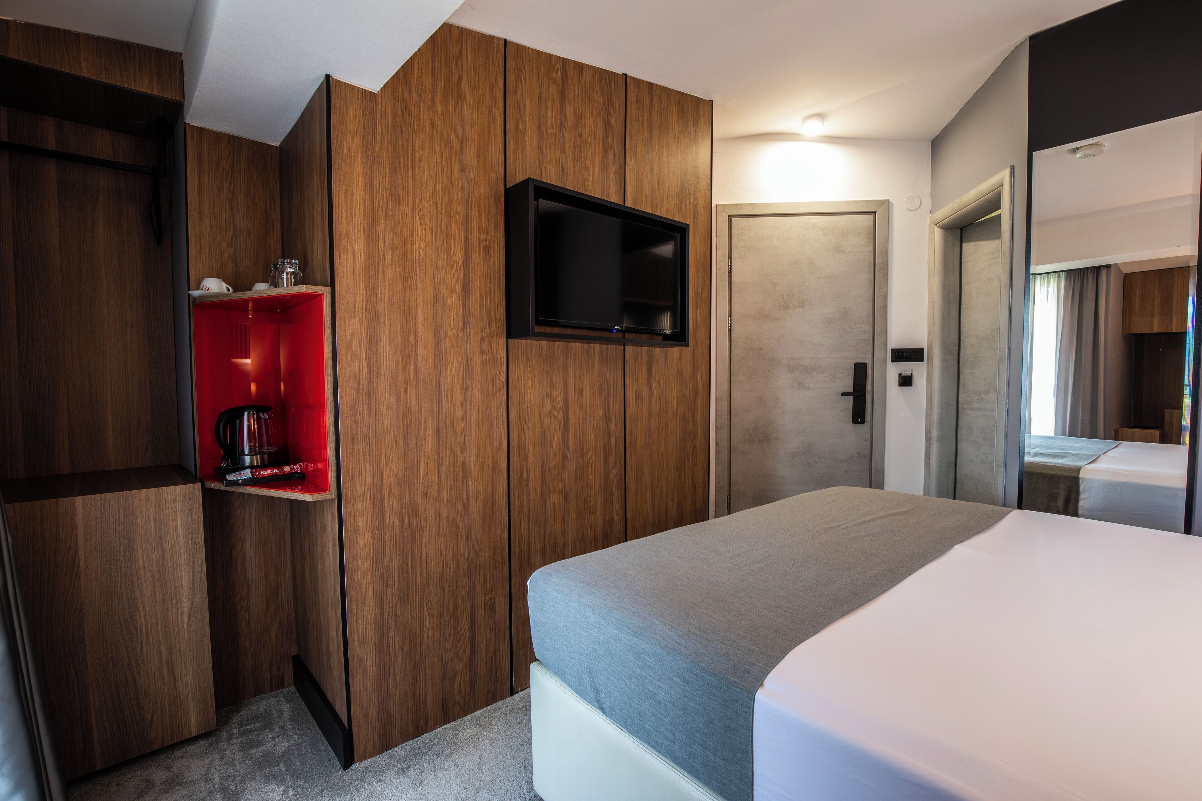 Quarto de hotel com uma cama, uma televisão fixada na parede e uma estante vermelha com uma cafeteira.