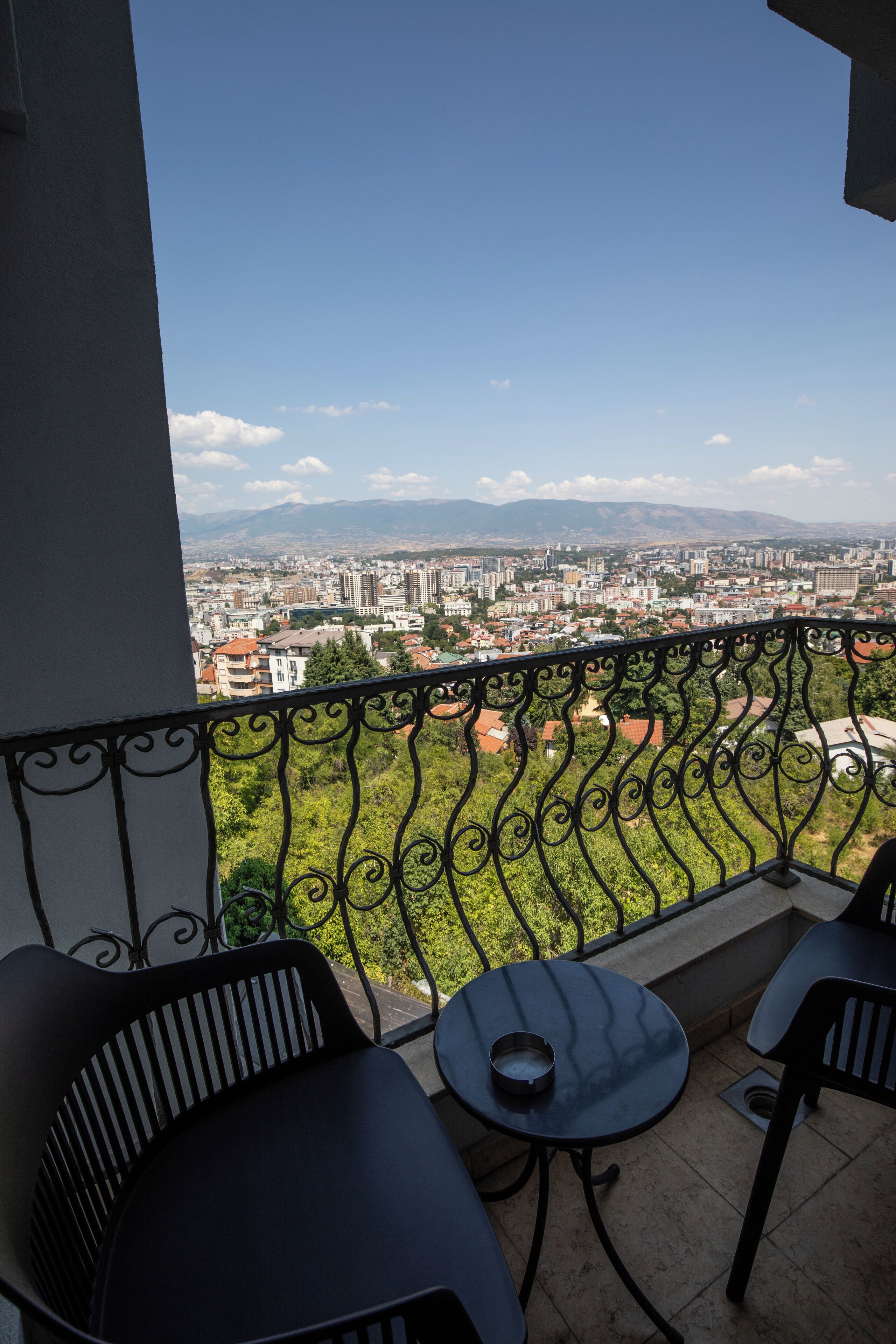 A balcony with a view of a cityscape, including buildings and mountains. There are two chairs and a small table on the balcony.