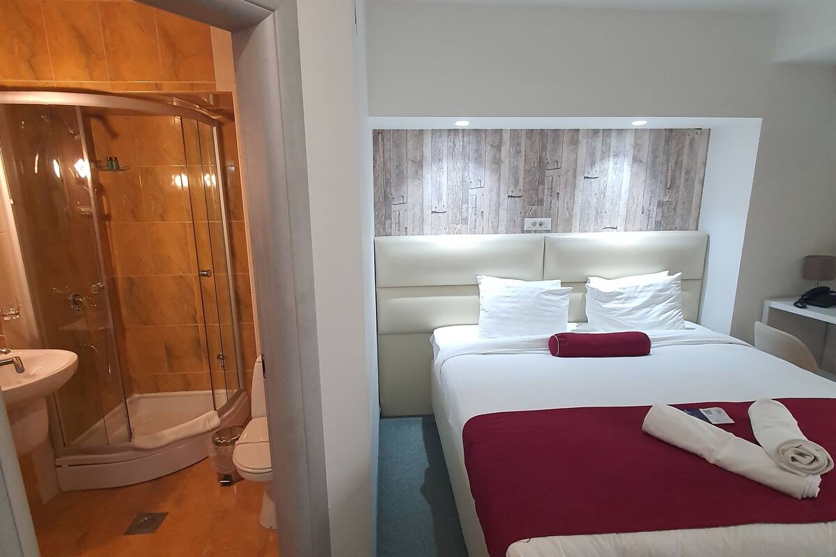 A hotel room with a shower, a bed with a red blanket, and a headboard with a wooden panel design.