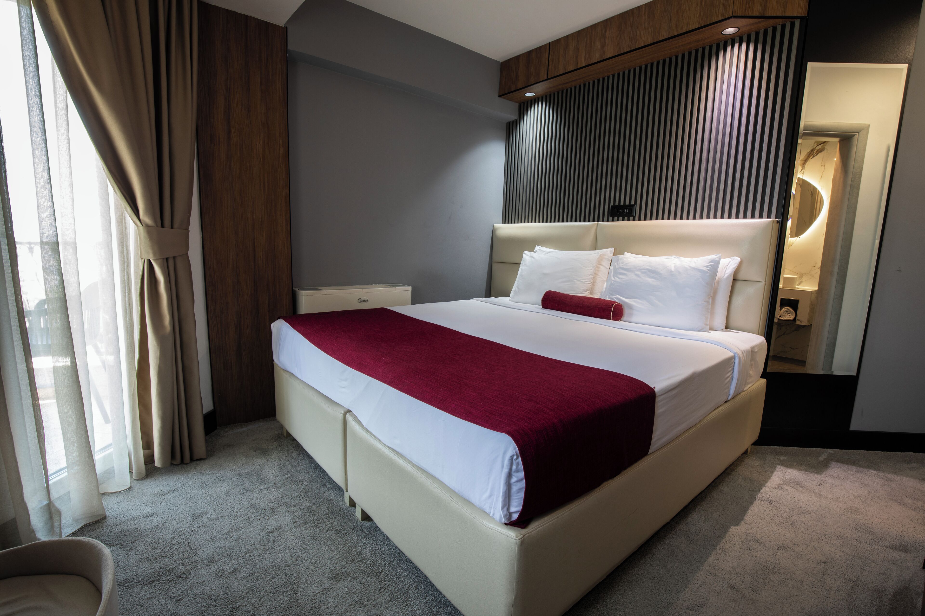 A modern hotel room with a large bed, a bedside table, a mirror, and a view of the outdoors.