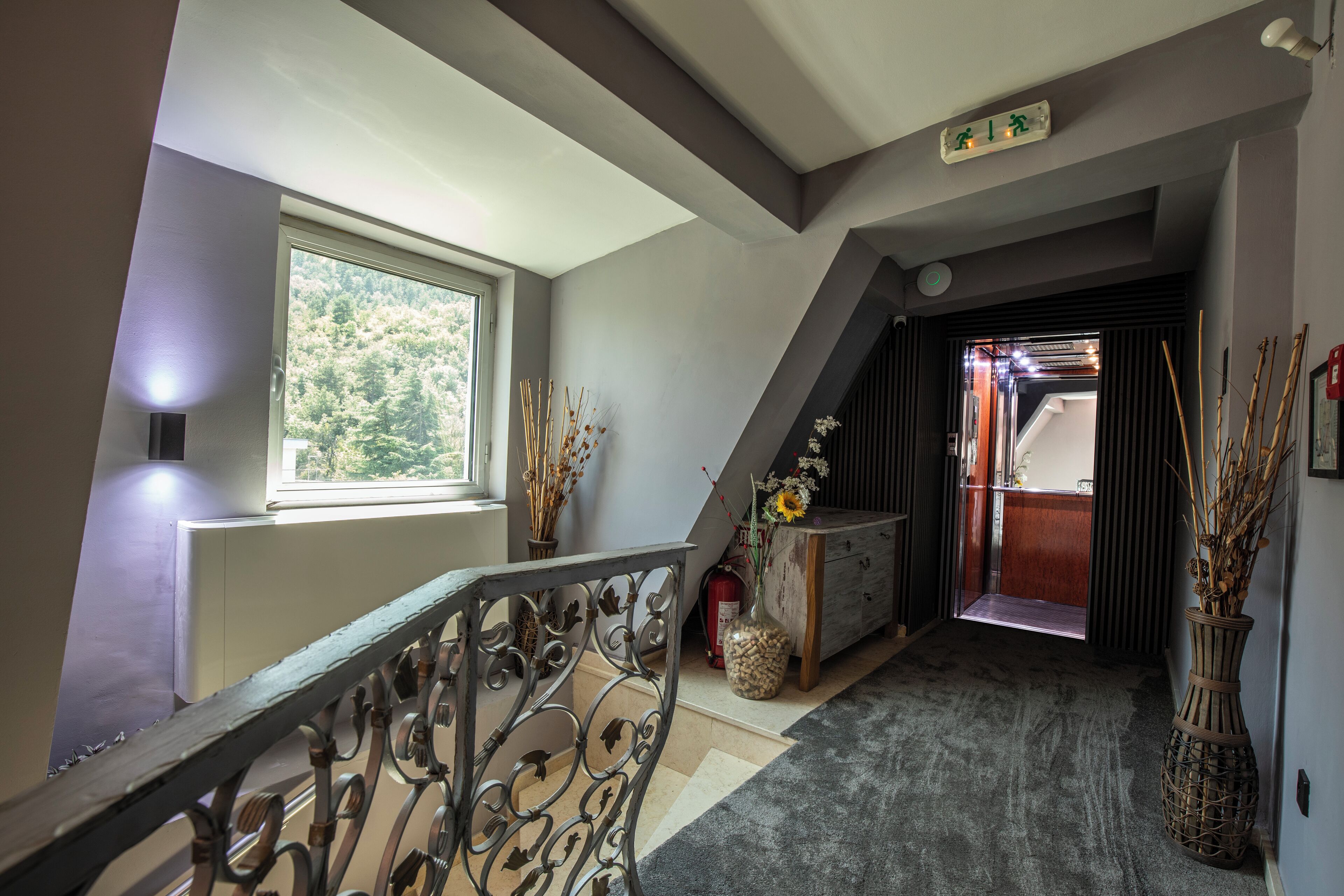 A hallway with a staircase, a window with a view of trees, a wooden cabinet, and a vase with dried plants.