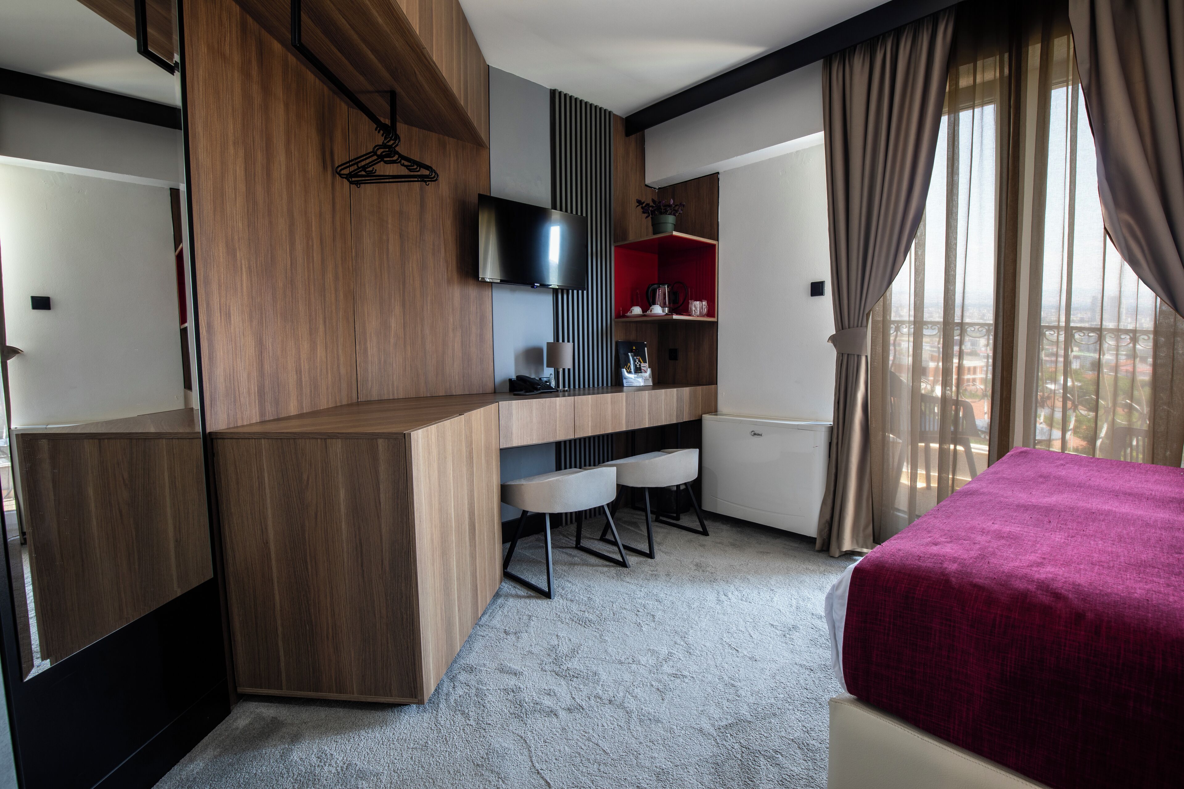 Una habitación de hotel moderna con un escritorio de madera, un televisor, una cama con cubierta roja y vistas a la ciudad.