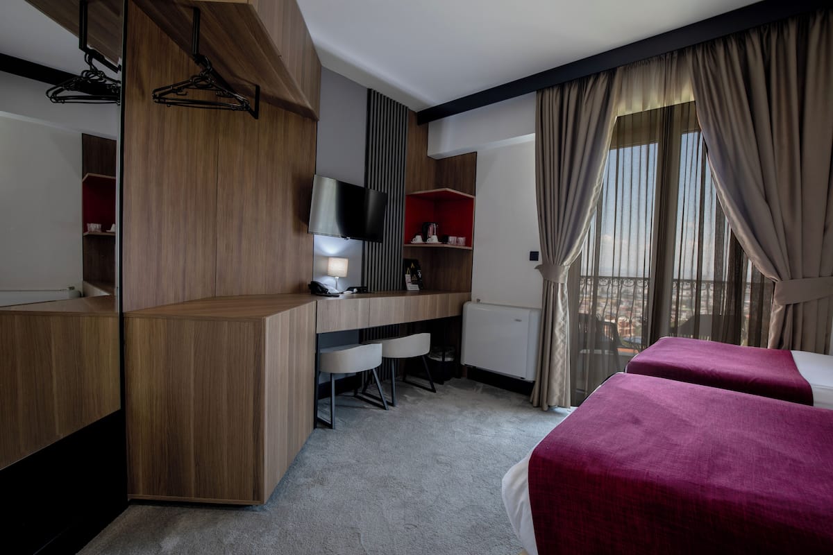 A hotel room with a wooden desk, two chairs, a bed with red bedding, a TV, and a window with curtains.