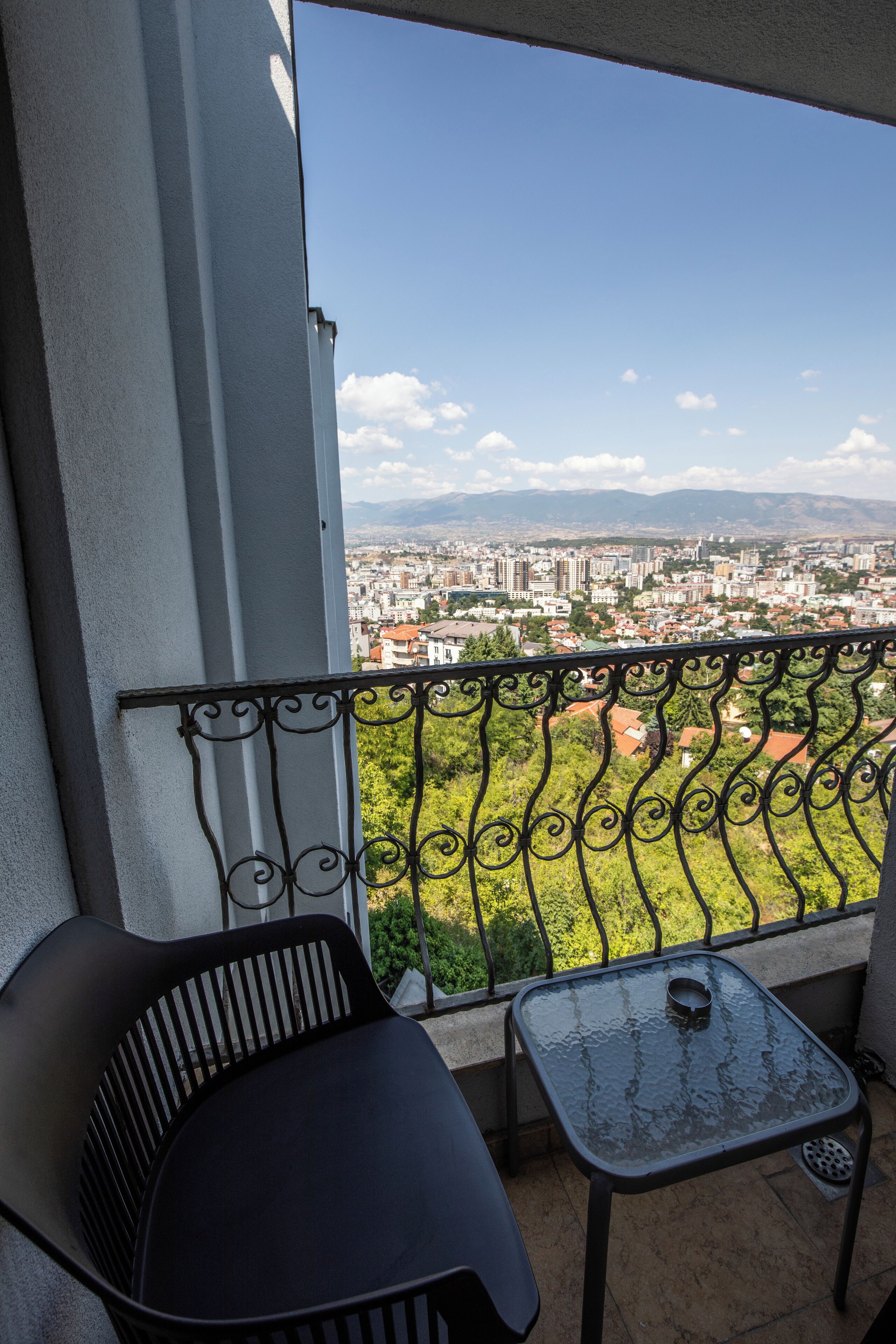 A balcony with a view of a cityscape, a black metal chair, and a small table with a cup.