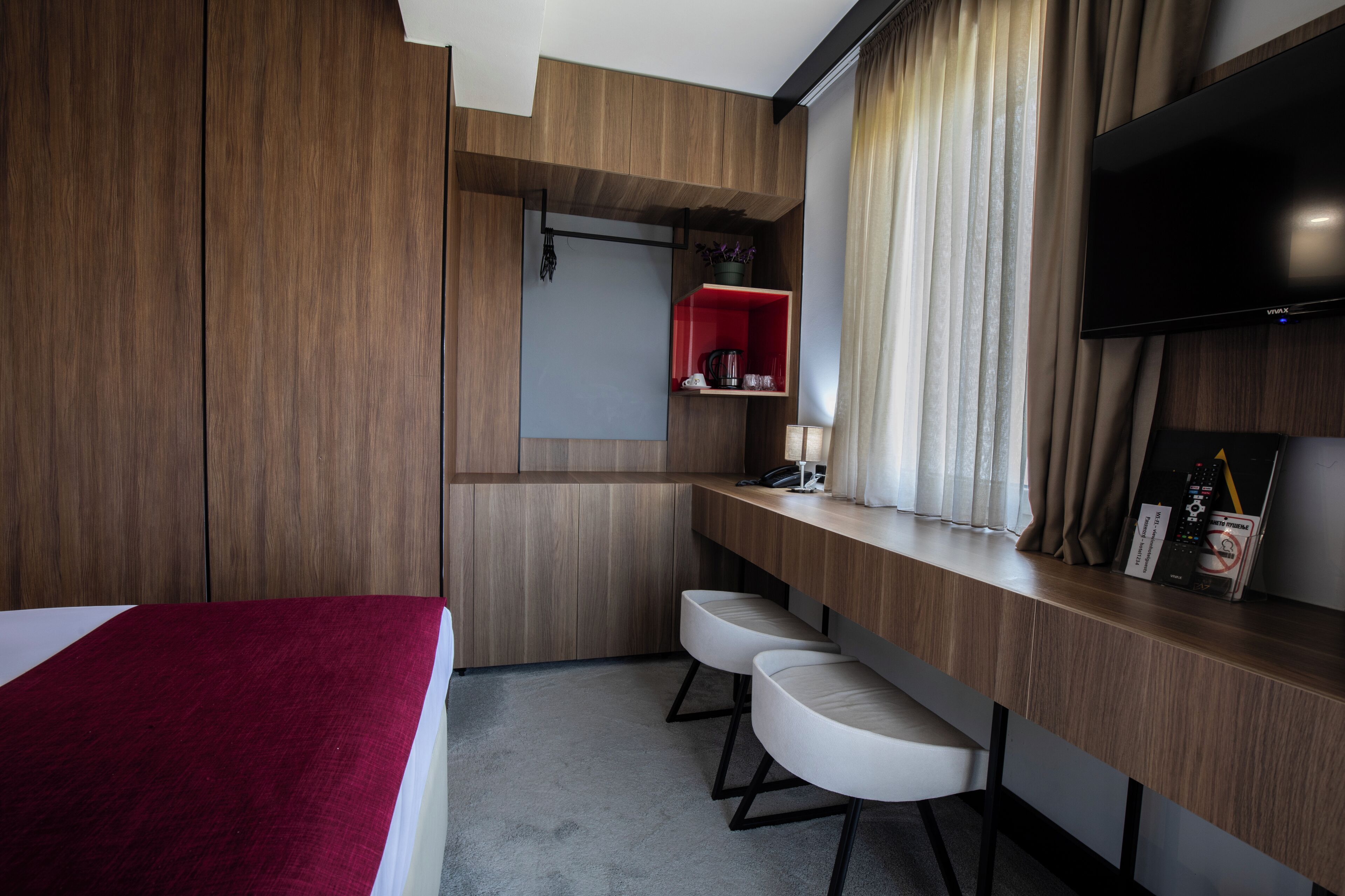 Habitación de hotel con cama, escritorio con dos sillas, televisor y estante rojo con cafetera.