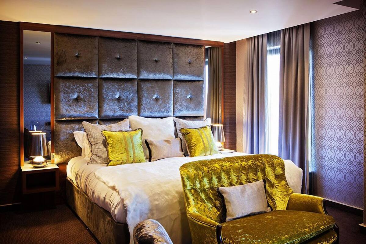 Een hotelkamer met een groot bed, een groene bekleden fauteuil en een nachtkastje met een lamp.