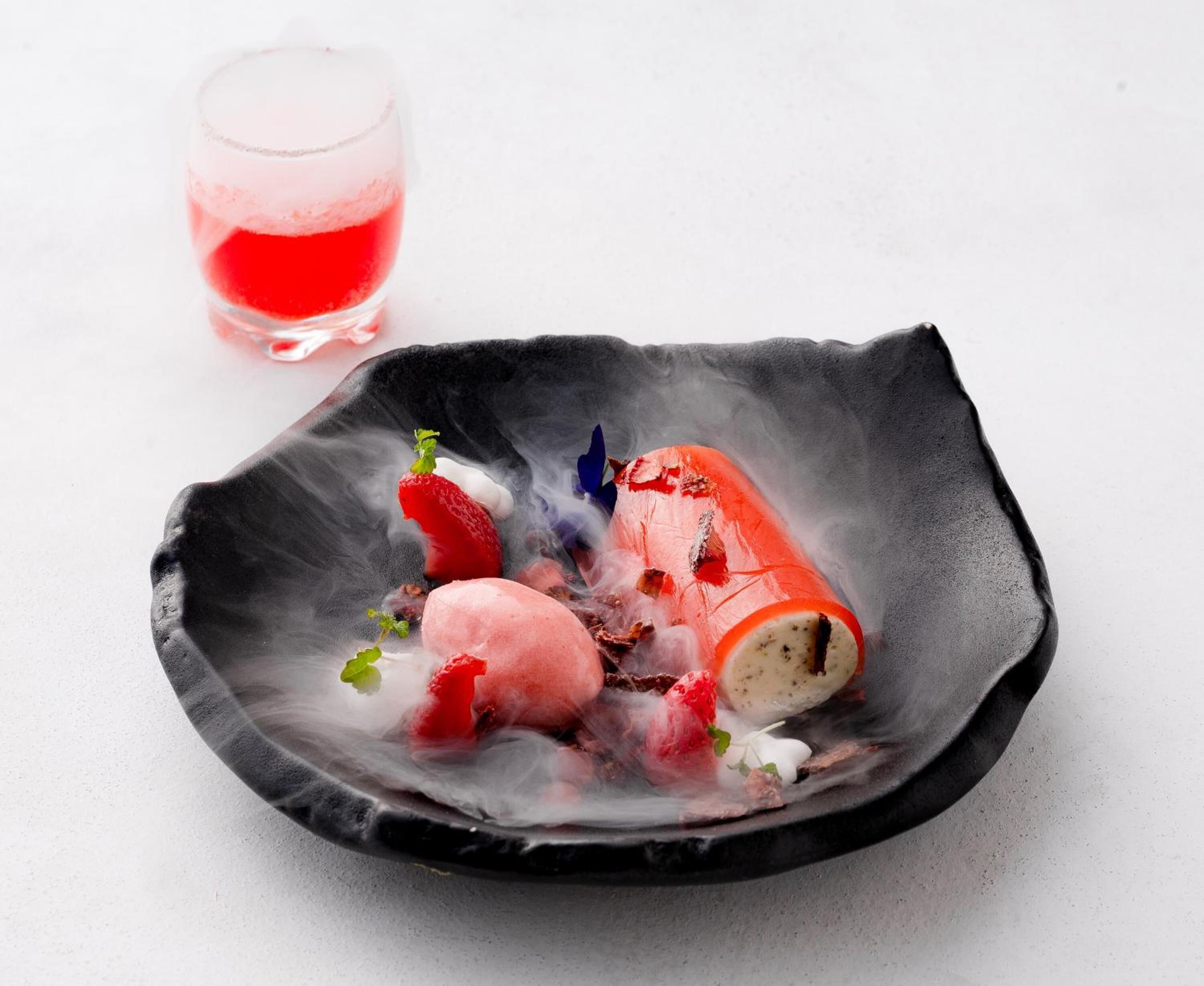 A dessert presentation on a black plate with ice cream and berries, accompanied by a glass of red beverage.