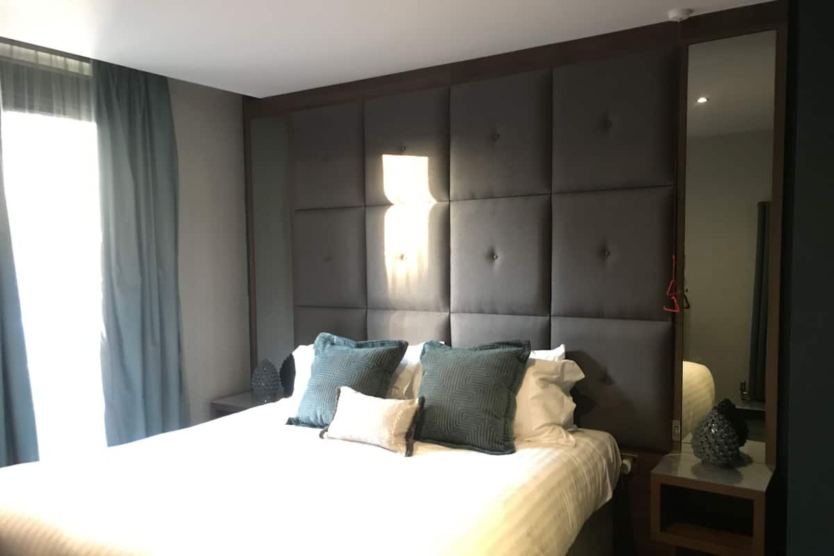 Een slaapkamer met een groot bed, een hoofdeinde met een lamp, een nachtkastje met een lamp, en een spiegel.