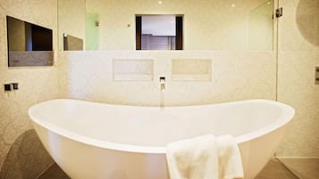 A modern bathroom with a freestanding bathtub, a large mirror, and a view of a room with a bed and a window.