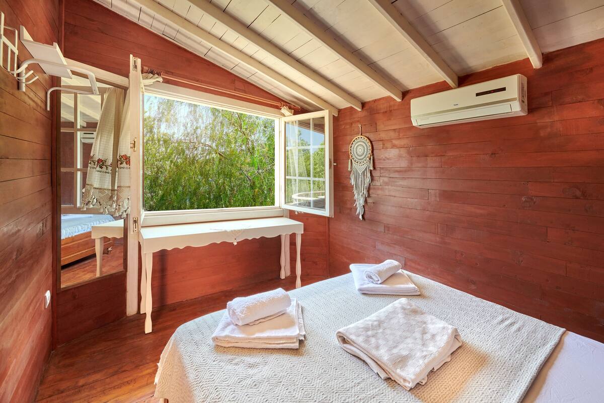 A wooden room with a bed, a window, a bench, and a wall-mounted air conditioner.