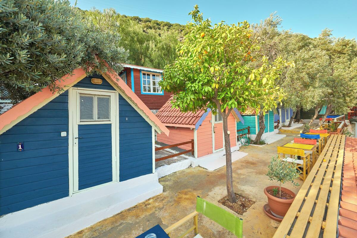 A row of colorful wooden cabins with a small tree in the center, a wooden bench, and a pathway leading to a door.