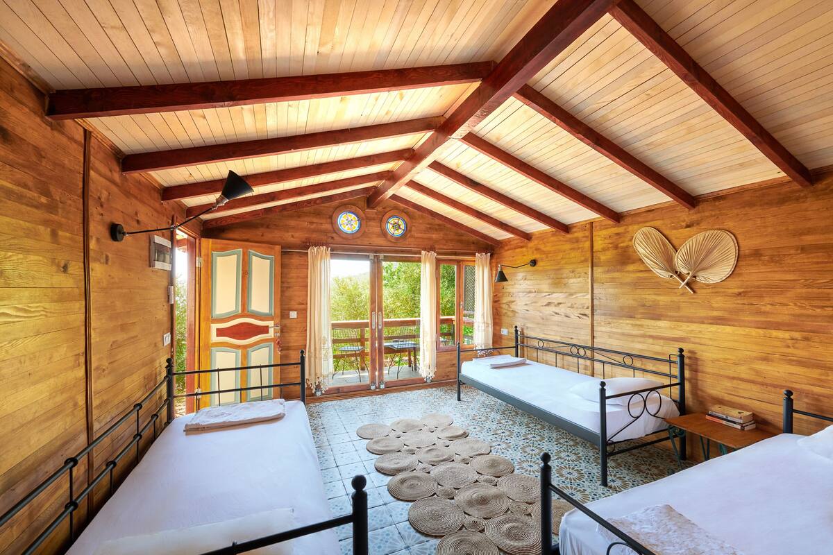 A wooden cabin with two beds, a large window, and a fan on the wall.