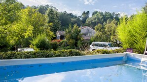 A swimming pool with a blue and white border, surrounded by lush greenery and houses in the background. - La Ribeyrette (Chamborigaud)