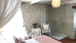 A bedroom with a bed, a chair, a table with a vase of flowers, a glass shower, and a hanging light fixture. - La Ribeyrette (Chamborigaud)