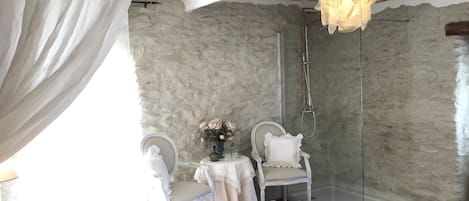 A bedroom with a bed, a chair, a table with a vase of flowers, a glass shower, and a hanging light fixture.