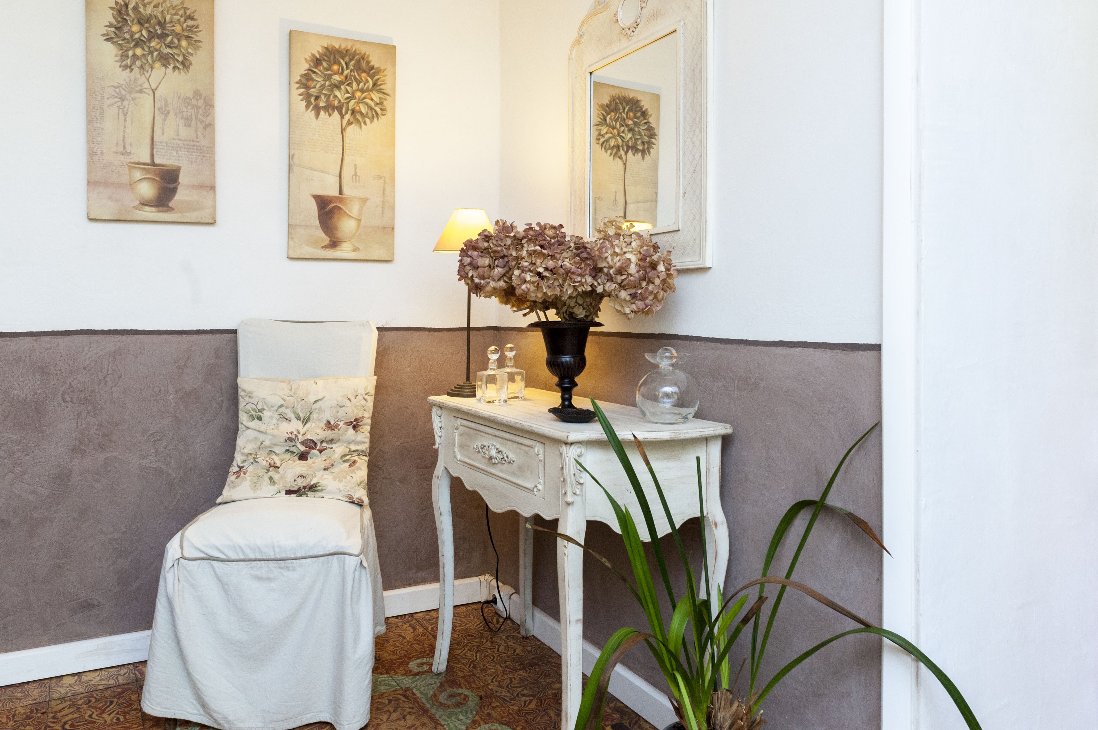Een hoekkamer met een witte stoel, een klein wit tafeltje met een lamp en een bloemenvaas. Twee schilderijen hangen aan de muur en er is een spiegel boven het tafeltje.