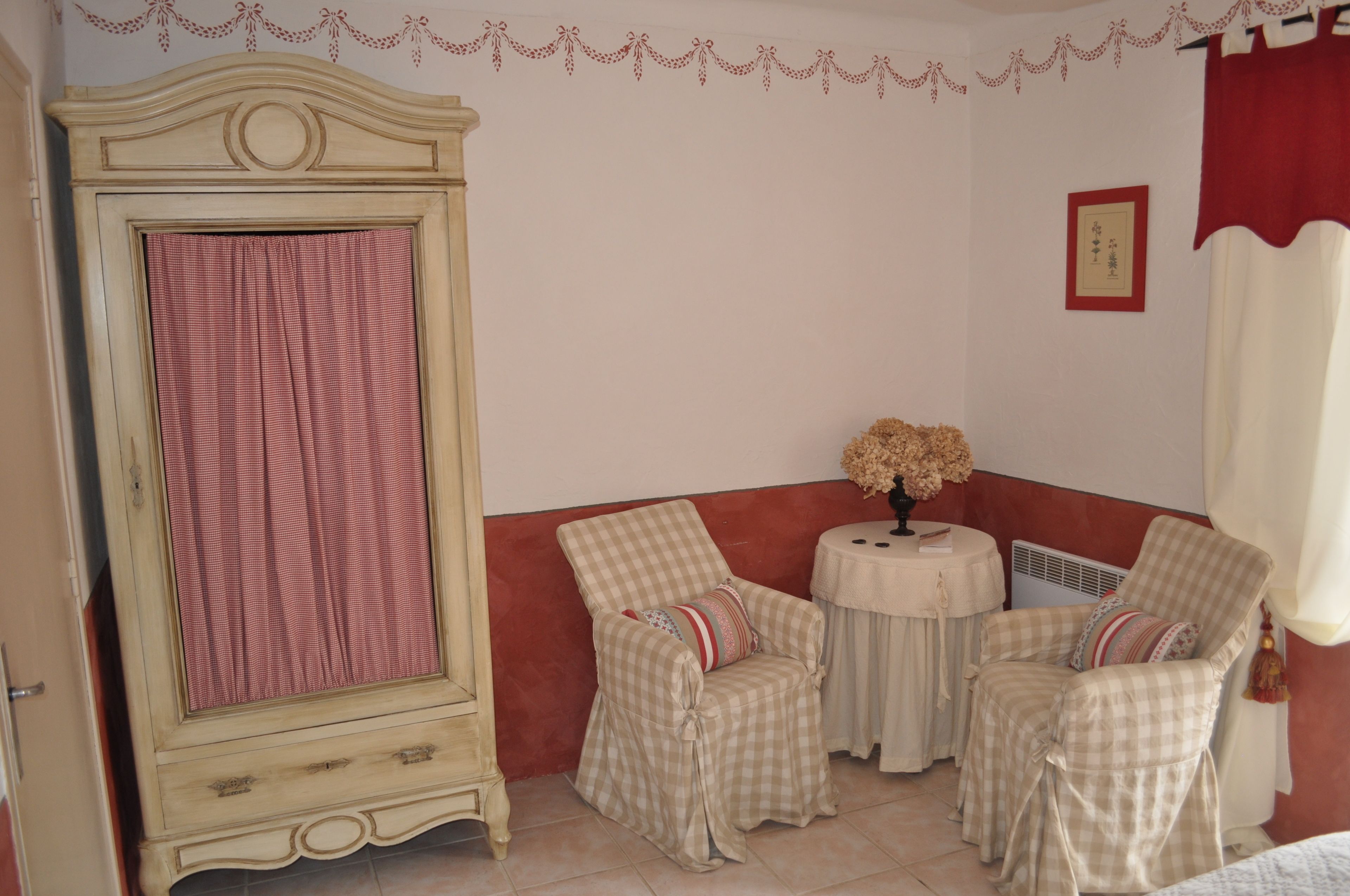 A room with a large wardrobe, two checkered chairs, a small round table with a vase, and a red curtain.