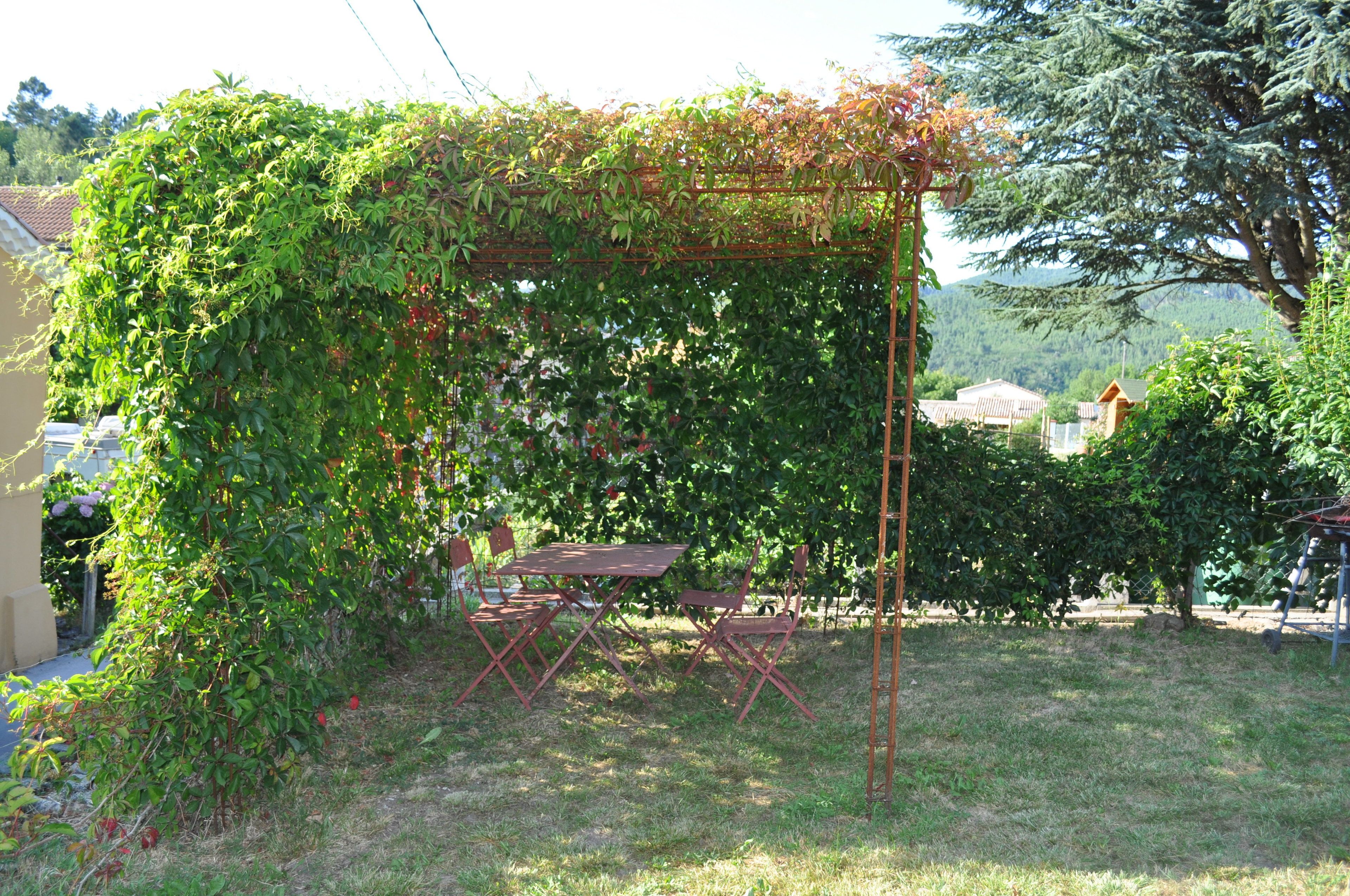 Um jardim com pérgola coberta por trepadeiras, uma mesa e cadeiras.