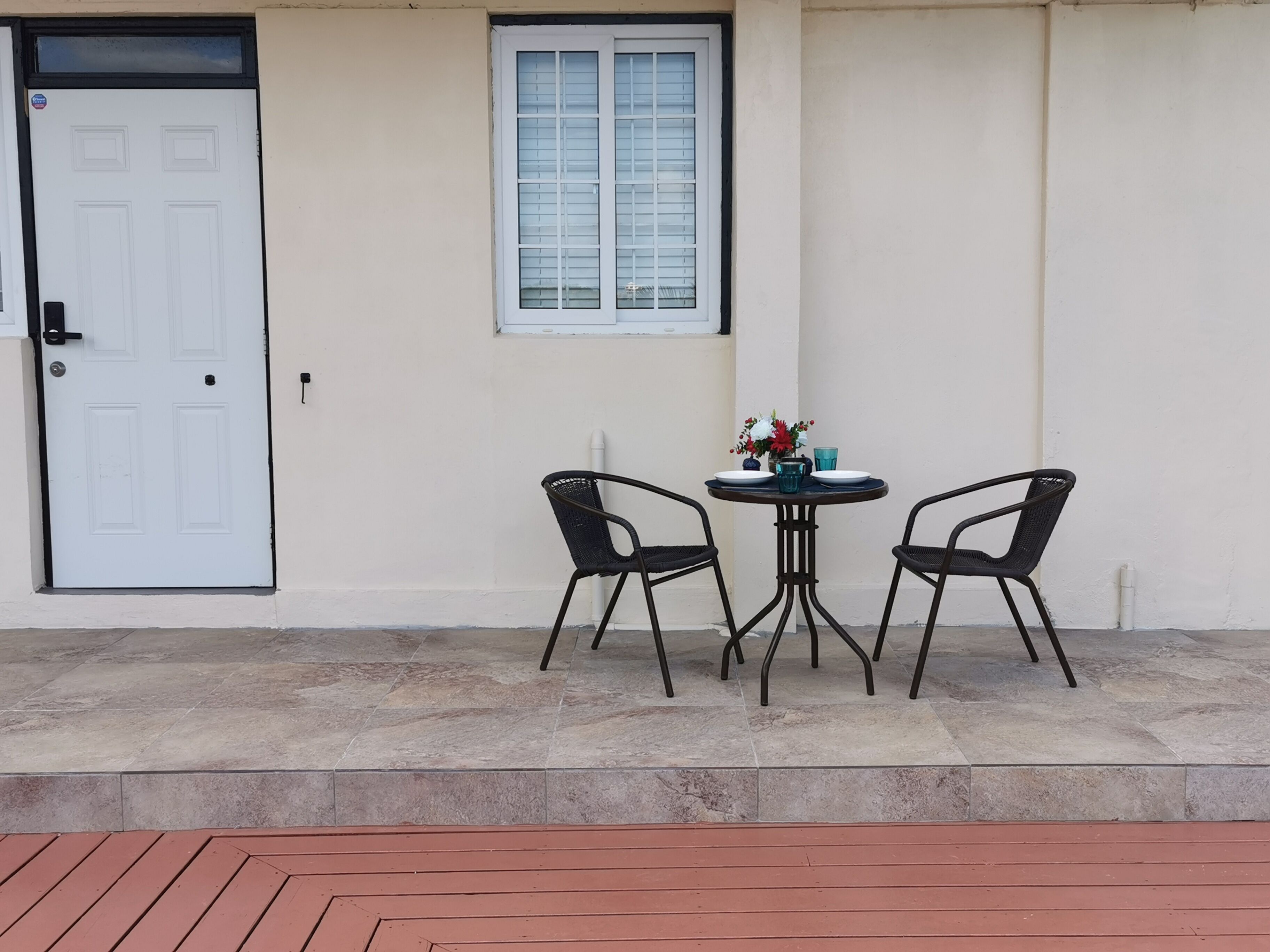 A small outdoor seating area with a round table and two chairs, a white door, and a window with blinds.