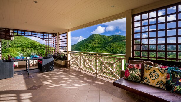 A covered balcony with a view of lush green hills, a dining area with a table set for two, and a seating area with patterned cushions. - Flamboyant Villa (Gros Islet)