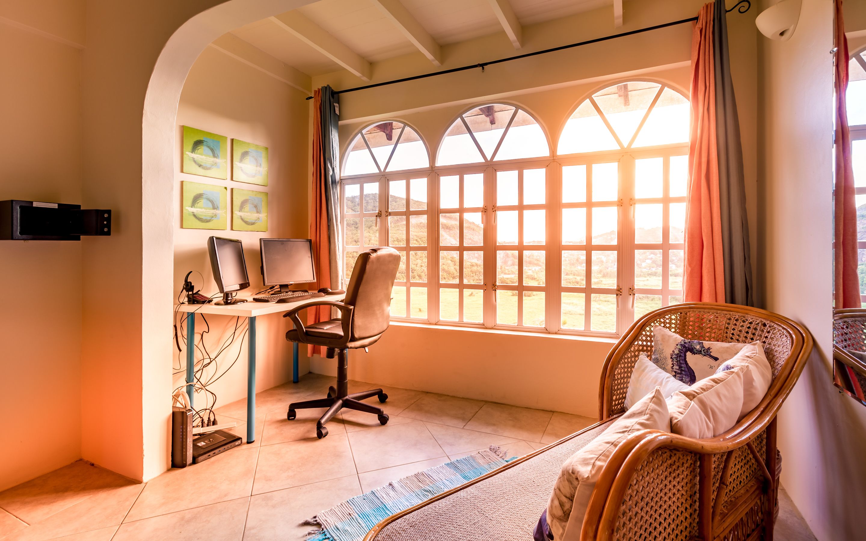 A room with a computer desk, two monitors, a chair, a wicker chair with pillows, and a large window with curtains.