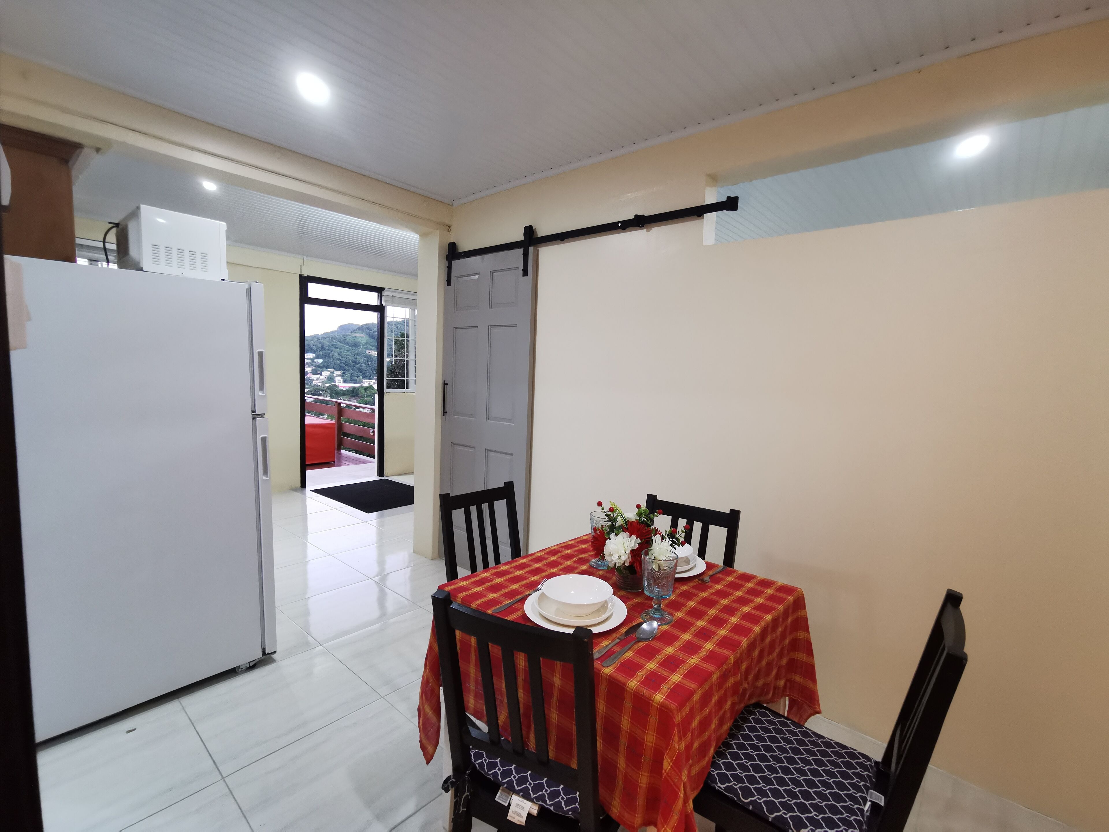 Un comedor con una mesa lista para dos, un refrigerador y una puerta que da a un balcón con vistas a las colinas.