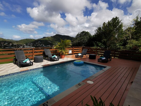 A pool area with lounge chairs and a wooden deck, surrounded by greenery and a mountainous backdrop.