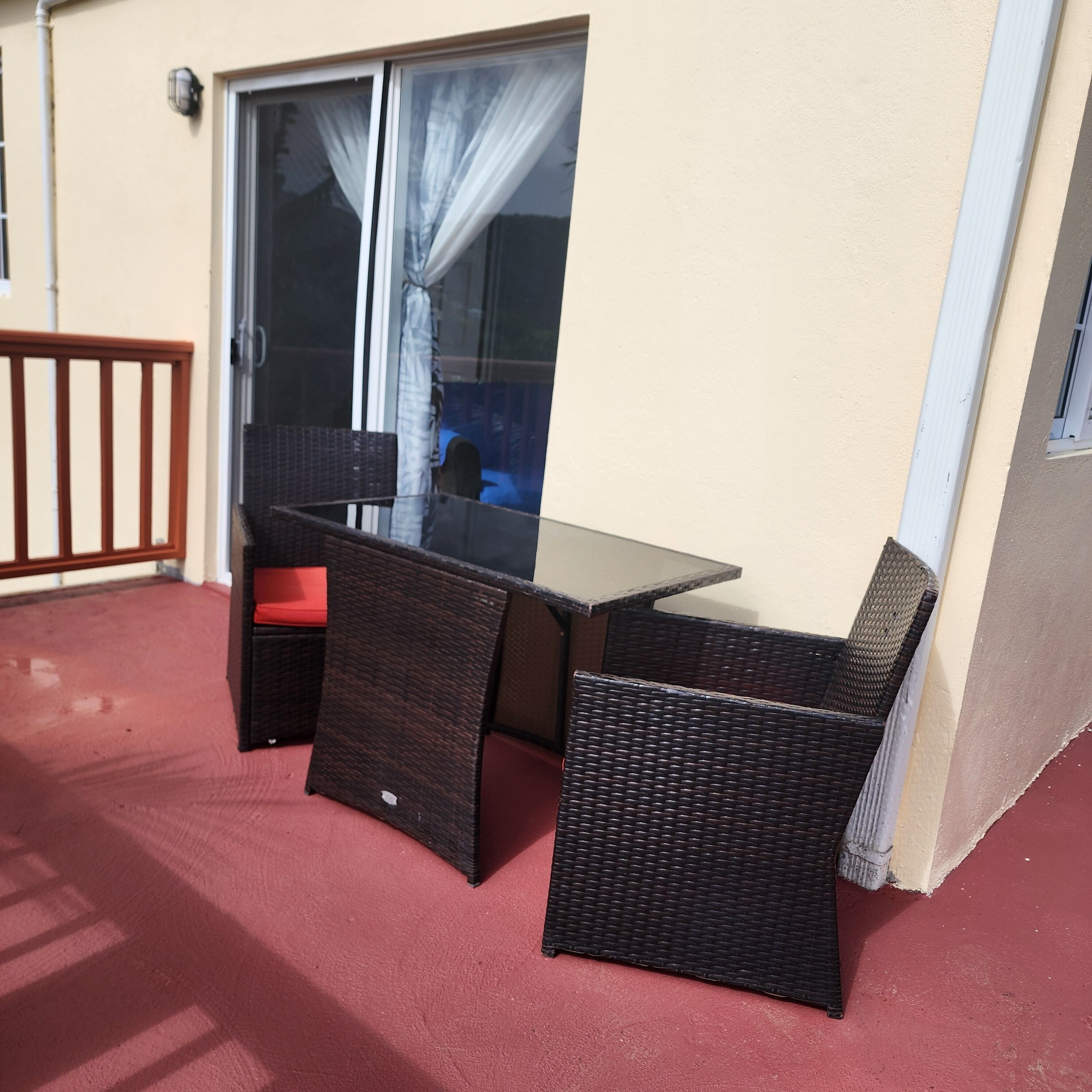 Un balcon avec une table en verre et des chaises en osier, une porte-fenêtre coulissante et un tapis rouge.