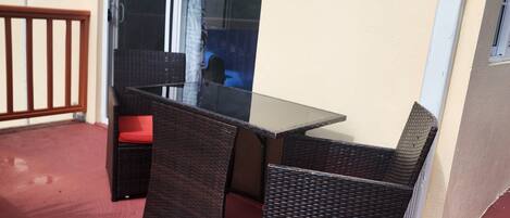 Un balcon avec une table en verre et des chaises en osier, une porte-fenêtre coulissante et un tapis rouge.