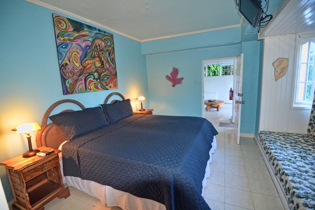 Un dormitorio con cama, mesitas de noche, un cuadro y una puerta que da a otra habitación.