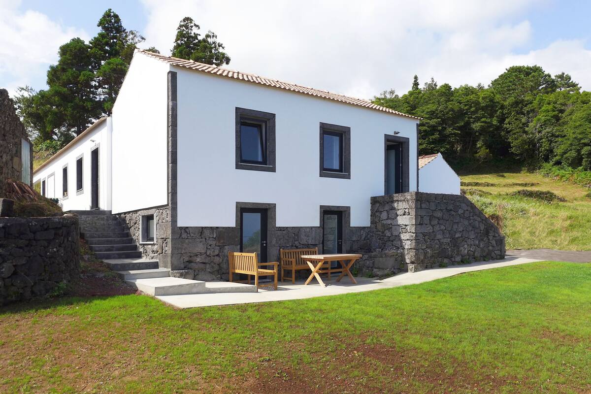 A two-story house with a stone foundation, white exterior walls, and a tiled roof. There is a small patio with a table and chairs.