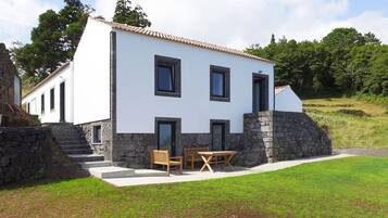 A two-story house with a stone foundation, white exterior walls, and a tiled roof. There is a small patio with a table and chairs.