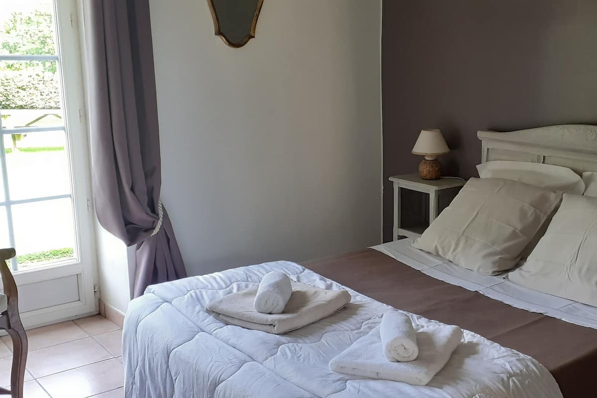 A neatly made bed with white linens and towels, a bedside table with a lamp, and a view of the outdoors through a window with curtains.