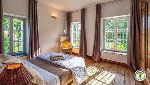A bedroom with two beds, a wicker chair, a lamp, and large windows with curtains. - Gîtes Normands de charme Les Châtaigniers (Bretteville-du-Grand Caux)