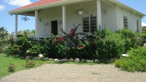 A single-story house with a red roof, a covered porch, and a garden with various plants and flowers. - Friars Hill Cottage (St. John's)